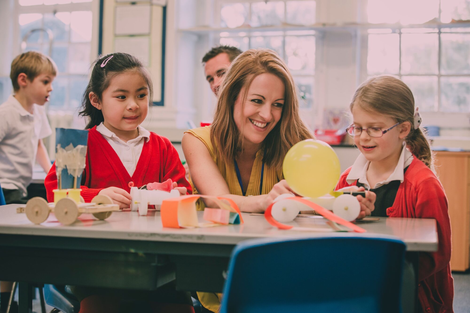 a young teacher smiling as she works with two young girls in primary school to create a creative project