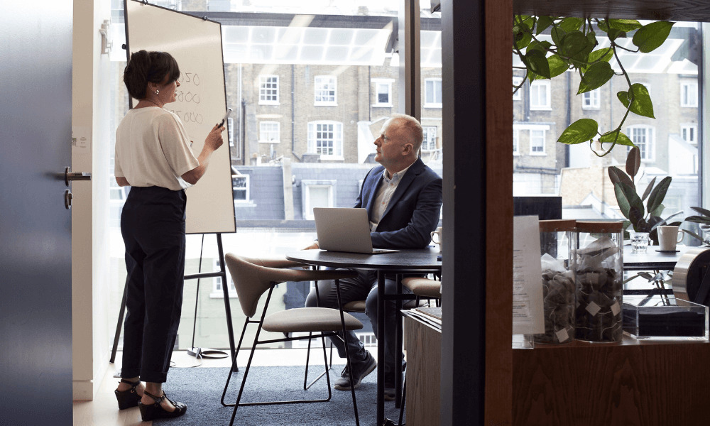 Two people in an office, the first is a man in a suit sat down, the next is a woman stood up by a whiteboard pointing at numbers