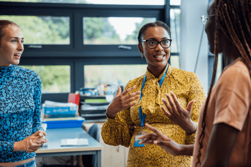 A young black woman in a yellow shirt and glasses with her hair tied back in two plaits and a lanyard on, looking at another colleague as though she is explaining something
