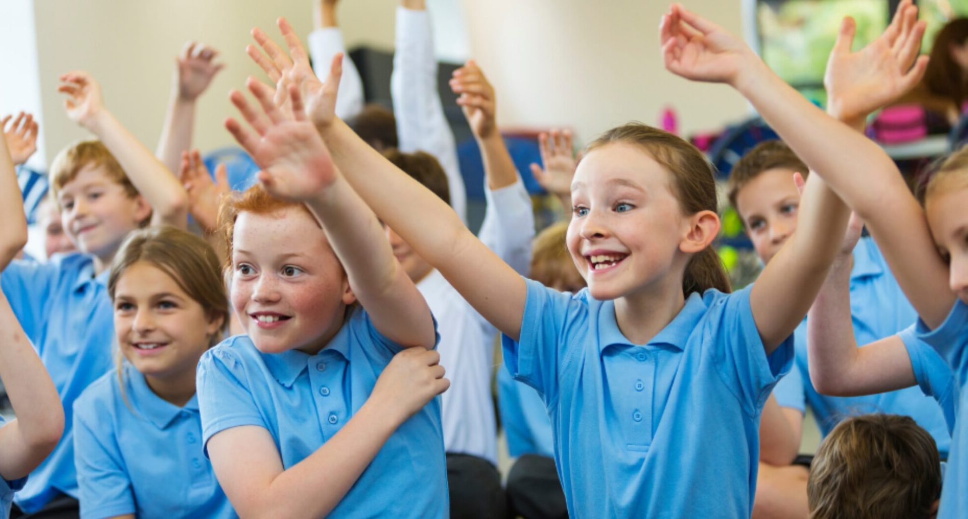young primary school children in blue uniform looking excited and puting their hands up