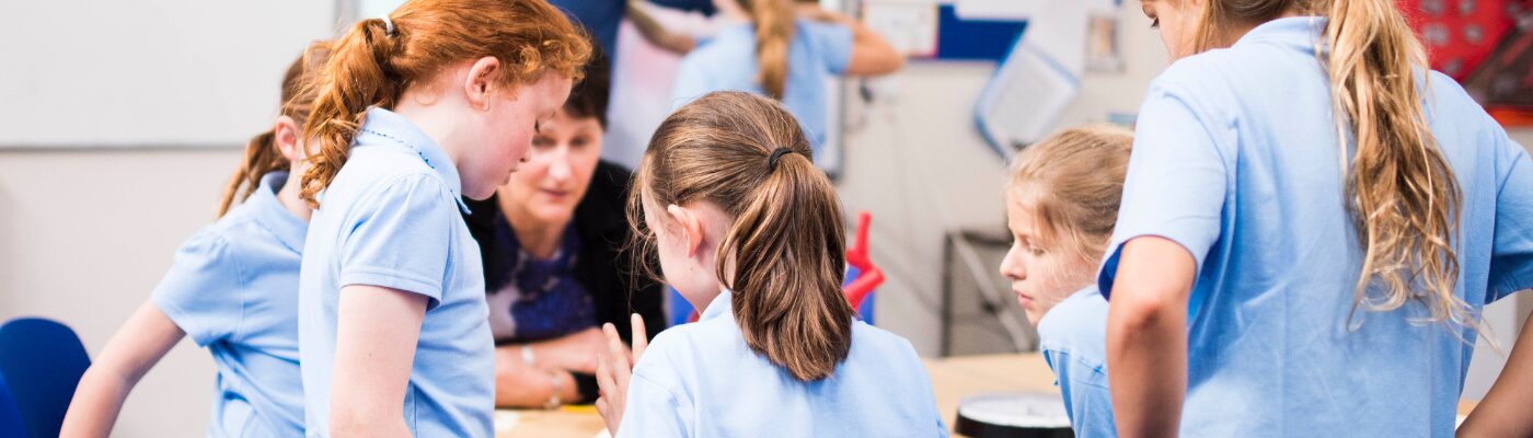 Young children in school uniform working on something with a teacher in the blurred distance